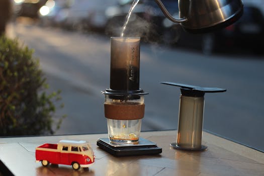 Aeropress coffee setup on table with hot water pouring from kettle and toy car nearby.