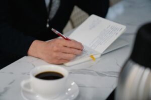 Businessman's hand writing notes in a journal with black coffee beside, indoors setting.